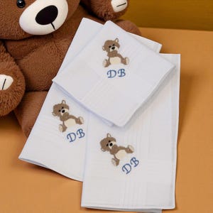 May include: White handkerchiefs with a teddy bear embroidery and the initials "DB" in blue. The handkerchiefs are folded and stacked, with a brown teddy bear in the background. The handkerchiefs have a striped pattern.