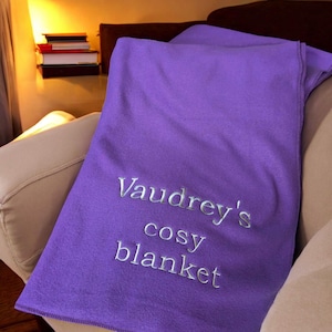 May include: A solid purple fleece blanket featuring silver embroidered text that reads Vaudrey's cosy blanket. The fabric has a smooth, soft texture and is draped over a piece of furniture, showcasing the personalized lettering clearly.