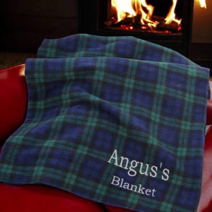 May include: A blue and green tartan blanket with the text "Angus's Blanket" in white. The blanket is folded on a red surface, with a fireplace in the background.