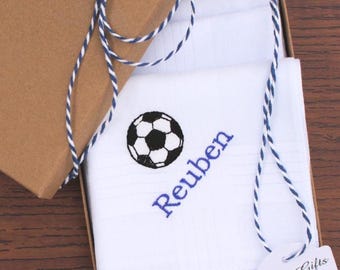 Könnte beinhalten: Ein weißes Baumwolltaschentuch mit einem gestickten schwarz-weißen Fußball und dem Namen Reuben in blauem Garn. Das Taschentuch liegt gefaltet in einer braunen Pappschachtel mit blau-weißer Schnur und einem Geschenkanhänger.