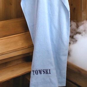 May include: A light blue towel with the word "TOWSKI" embroidered in navy blue. The towel is hanging in a wooden sauna, with steam visible in the background. The towel appears to be made of a soft, absorbent material.