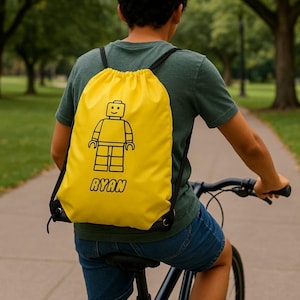 Op de afbeelding: Een gele trekkoordrugzak met een zwarte Lego-figuur en de naam "RYAN". De rugzak, met zwarte trekkoorden, wordt gedragen door iemand die op een fiets rijdt. De achtergrond is een park.
