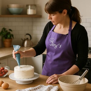 May include: A purple apron featuring a white chef hat icon and the text Joy's embroidered on the chest. The apron has adjustable neck straps and is worn over a black long-sleeved shirt while decorating a round cake with a blue piping bag.