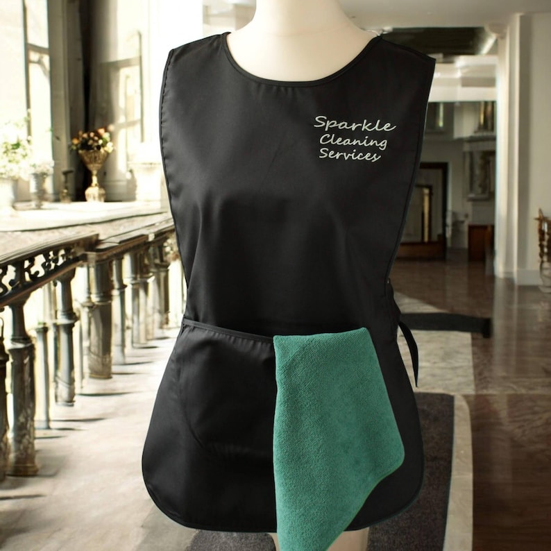 Puede incluir: Delantal de limpieza negro con un bolsillo grande y un pa&ntilde;o de limpieza verde. El delantal tiene el texto "Sparkle Cleaning Services" bordado en la parte delantera.