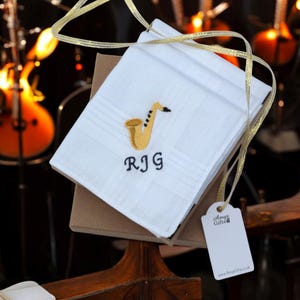 May include: A white handkerchief with a gold embroidered saxophone and the name "RJG" in black embroidery. The handkerchief is folded and tied with a gold ribbon. It is sitting on a brown box with a white tag that says "Amy's Gifts".