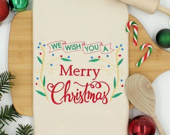 May include: A cream-colored Christmas kitchen towel with embroidered text reading "We Wish You a Merry Christmas." The design includes red, green, and blue accents. Candy canes and ornaments are arranged around the towel on a wooden cutting board.
