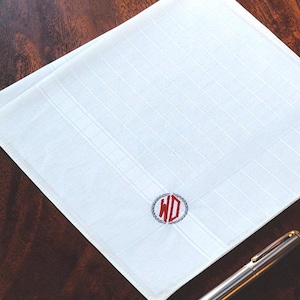 May include: A white square pocket square featuring a subtle grid pattern and a circular embroidered emblem in the corner. The emblem displays the red letters WD enclosed within a grey laurel wreath design. A metallic pen is visible at the bottom.