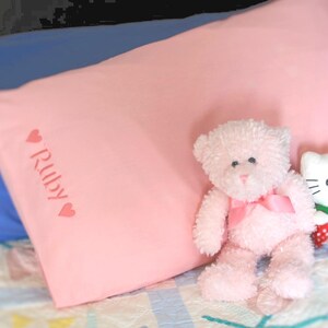 May include: A light pink pillow with the name "Ruby" printed in a cursive font, flanked by heart symbols. A pink teddy bear with a pink bow sits next to the pillow. The teddy bear has black eyes and a pink nose.