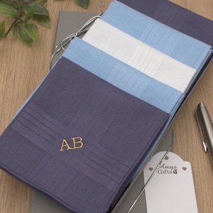 May include: A set of folded square handkerchiefs in shades of navy blue, light blue, and white, stacked inside a grey gift box. The top navy blue handkerchief features the embroidered gold initials AB. A white gift tag reads Amys Gifts.