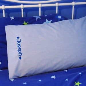 May include: A light blue pillow with the name "Joseph" embroidered in blue, accented with stars. The pillow rests on a blue patterned bedsheet with white stars. The bed frame is white.