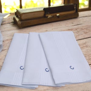 Op de afbeelding: Drie witte zakdoeken met een blauw geborduurd "C" liggen op een houten oppervlak. Elke zakdoek heeft verticale en horizontale stiklijnen. De achtergrond toont een raam en een houten doos.