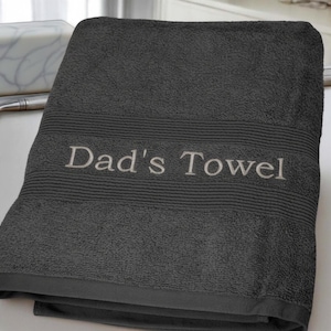 May include: A charcoal grey bath towel with the embroidered text "Dad's Towel" in a light grey colour. The towel has a textured surface and a ribbed border design.