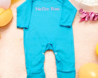 May include: A turquoise baby onesie with long sleeves and attached feet. The text "Nellie Rae" is printed in pink across the chest. The onesie has snap closures at the crotch. Balloons and a cake are in the background.
