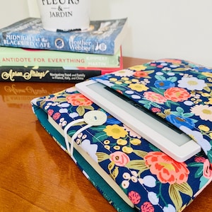 May include: A navy blue floral print e-reader case with a white e-reader inside. The case has a button closure and is on top of a stack of books. A small white pot with the text "FLEURS & JARDIN" is on top of the books.