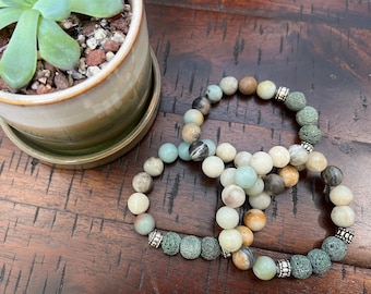 May include: Three beaded bracelets with green, brown, and white stones. The bracelets have silver accents and are stacked on a wooden surface.