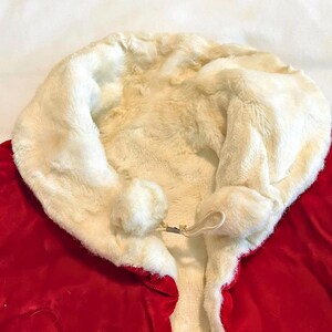 May include: A red Santa Claus costume with a white faux fur trim and a matching white fur-lined hood. The hood has two white pom-poms and a metal clasp closure. The costume is made of a soft, plush material.