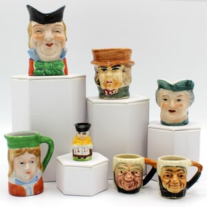 Puede incluir: Una colección de siete pequeñas tazas de cerámica con caras pintadas. Las tazas son de varios colores, incluyendo verde, marrón y azul. Las tazas se muestran en pedestales blancos.