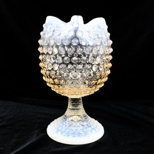 Może przedstawiać: Opalizujący szklany wazon z hobnail, z rozkloszowaną górą i podstawą na cokole. Tekstura hobnail pokrywa korpus wazonu, tworząc wygląd bąbelkowy. Wazon ma miękką, świecącą jakość, z gradientem koloru od białego do żółtego.