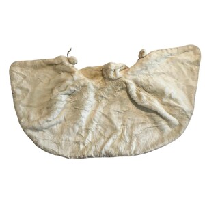 May include: A cream-colored, textured sculpture with a rounded shape. The artwork features three figures with outstretched arms, resembling wings. The piece has a soft, organic appearance, with subtle variations in tone and texture.