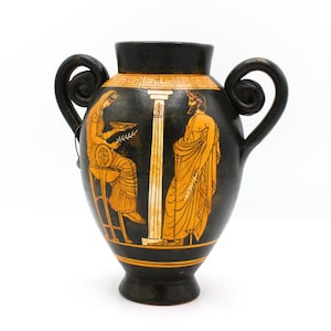 May include: Black ceramic vase with two curved handles. The vase features orange illustrations of figures and a column against a black background. The figures are in classical attire, and the vase has an orange band around the top and bottom.
