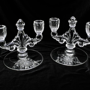 May include: A pair of clear glass candle holders with three candle cups each. The holders have a decorative, ornate design and a round base with an etched floral pattern.