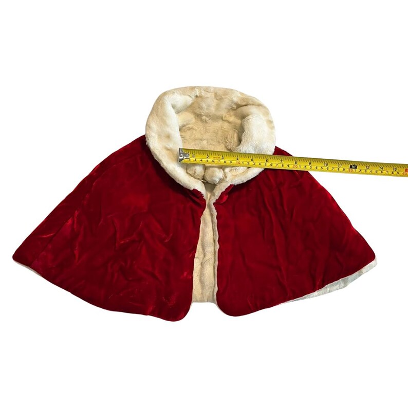 May include: A red velvet cape with a cream-colored faux fur collar and lining. The cape is measured with a tape measure, showing a width of approximately 16 inches.