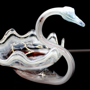 May include: A decorative glass swan bowl with a wavy rim. The swan's neck curves gracefully, and the bowl features white, brown, and clear glass. The swan's head is a light blue colour. The bowl is set against a black background.