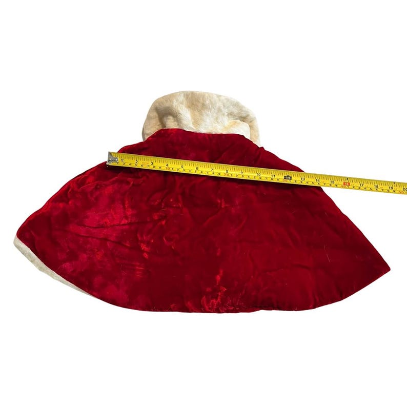 May include: A red velvet cape with a cream-colored faux fur collar and trim. The cape measures approximately 20 inches in length. The cape is a festive garment, suitable for holiday or theatrical use.