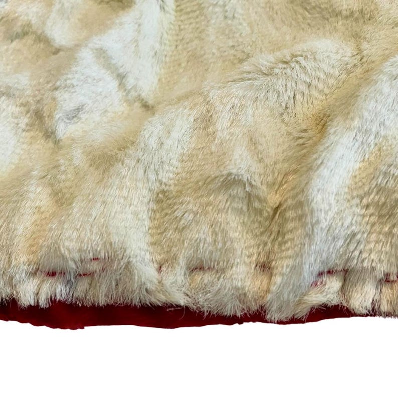 May include: Close-up of a plush, faux fur fabric in cream and tan hues. The fabric has a dense, textured pile, with visible stitching along the bottom edge where it meets a deep red fabric. The overall effect is soft and luxurious.