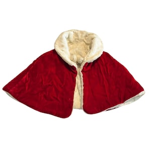 May include: A red velvet cape with a cream-colored faux fur collar and lining. The cape is open in the front and appears to be reversible. The garment is likely for a costume or festive occasion.