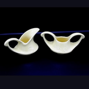 Op de afbeelding: Twee crèmekleurige keramische serveerschalen. Eén is een melkkan met een gebogen tuit en handvat. De andere is een suikerpot met twee handvatten. Beide hebben een wervelontwerp aan de voorkant.