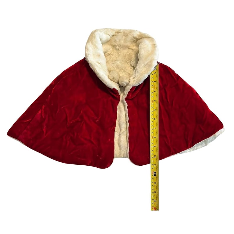 May include: A red velvet cape with a cream-colored faux fur collar and lining. The cape measures approximately 15 inches in length. The garment is open in the front and appears to be in good condition.