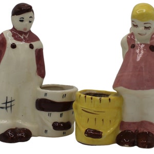 May include: A pair of vintage ceramic planter figurines. The figurines are a boy and a girl, each holding a small planter. The boy is wearing overalls and a red shirt, and the girl is wearing a pink dress. The planters are painted with brown and yellow stripes.