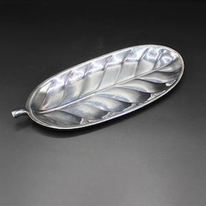 May include: A silver-colored leaf-shaped dish with detailed veining. The dish has a curved, elongated shape and a small stem at one end. The reflective surface suggests a metal material, possibly silver or a silver-toned alloy. The dish is designed for decorative or serving purposes.