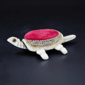 May include: A turtle-shaped pin cushion with a red velvet top and a decorative, ornate base. The turtle is white with gold accents and a small, dark gem for an eye. The cushion is approximately 4 inches long.