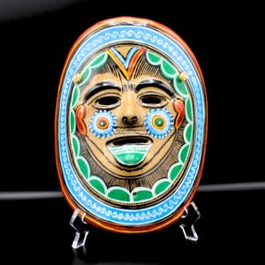 May include: Oval-shaped, hand-painted ceramic mask with a smiling face. The mask features a tan face with black outlines, blue flower accents, and a green mouth. A blue and green border surrounds the face, with red and yellow accents.