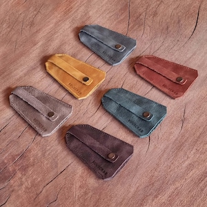 Może przedstawiać: Sześć skórzanych etui na klucze w różnych kolorach: szarym, musztardowym, czerwono-brązowym, morskim, brązowym i fioletowym. Każde etui ma zapięcie na zatrzask, a na każdym nadrukowano słowo "BOUSCRAB".