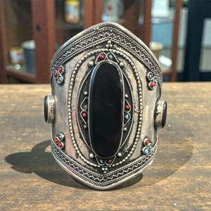 May include: A wide, silver-toned cuff bracelet with an oval black stone center. The bracelet features intricate patterns, red and blue accents, and two black stones on the sides. The design is ornate and detailed, showcasing a vintage or ethnic style.