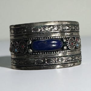 May include: A wide, silver-toned cuff bracelet with intricate etched patterns and a central, oval-shaped, deep blue stone. The bracelet features decorative elements in red and turquoise, adding pops of color to the design.