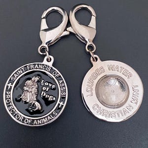 Puede incluir: Dos colgantes plateados con forma de chapa de perro. Uno presenta a San Francisco de As&iacute;s con el texto "Love of Dogs" y "Protector of Animals". El otro colgante tiene "Lourdes Water Christian Mint" alrededor de un centro transparente.