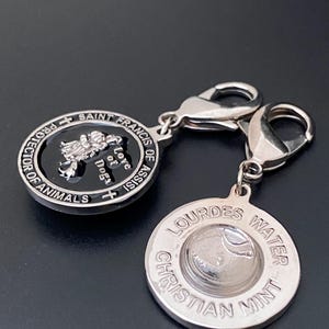 Puede incluir: Dos colgantes plateados. Un colgante presenta el texto "San Francisco de As&iacute;s, Protector de los Animales, Amor de Perros" alrededor de un centro negro con una imagen de perro. El otro colgante tiene "Lourdes Water Christian Mint" alrededor de un dise&ntilde;o central.