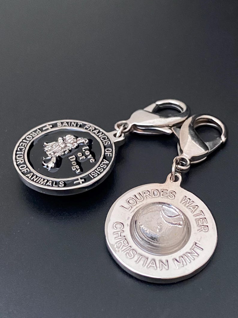 Puede incluir: Dos colgantes plateados. Un colgante presenta el texto "San Francisco de As&iacute;s, Protector de los Animales, Amor de Perros" alrededor de un centro negro con una imagen de perro. El otro colgante tiene "Lourdes Water Christian Mint" alrededor de un dise&ntilde;o central.