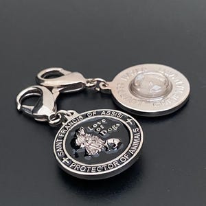 Puede incluir: Un charm de plata con dos colgantes conectados. Un colgante es redondo con el texto "WATER" y "LOUIS" alrededor de un centro transparente. El otro colgante es negro con el texto "SAINT FRANCIS OF ASSISI PROTECTOR OF ANIMALS" y "Love of Dogs".