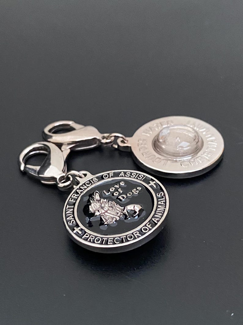 Puede incluir: Un charm de plata con dos colgantes conectados. Un colgante es redondo con el texto "WATER" y "LOUIS" alrededor de un centro transparente. El otro colgante es negro con el texto "SAINT FRANCIS OF ASSISI PROTECTOR OF ANIMALS" y "Love of Dogs".