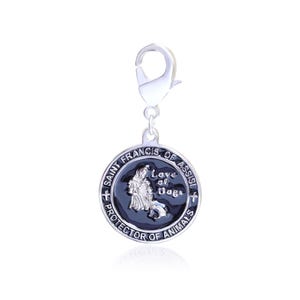 Puede incluir: Un dije plateado con un cierre de mosquet&oacute;n. El colgante circular presenta el texto "San Francisco de As&iacute;s, Protector de los Animales" y una imagen de San Francisco con un perro. El colgante es negro y plateado.