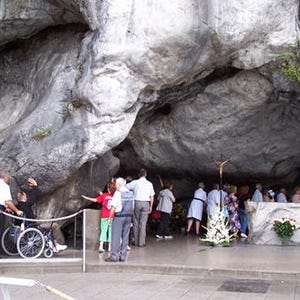 Puede incluir: Una gran cueva natural con un techo de roca gris y alto. Un grupo de personas se re&uacute;ne dentro de la cueva, algunas mirando hacia un peque&ntilde;o altar con un crucifijo. Hay una persona en silla de ruedas. Hay velas blancas a la derecha.