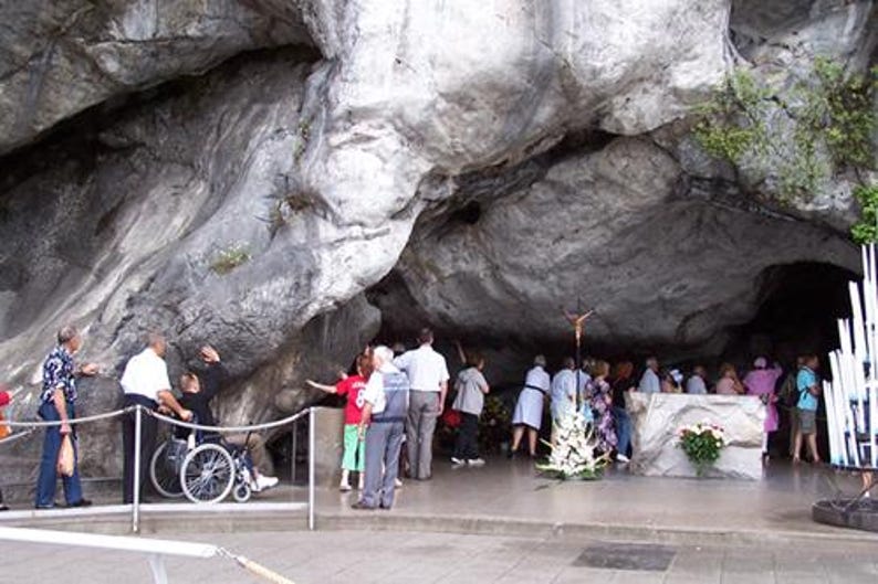 Puede incluir: Una gran cueva natural con un techo de roca gris y alto. Un grupo de personas se re&uacute;ne dentro de la cueva, algunas mirando hacia un peque&ntilde;o altar con un crucifijo. Hay una persona en silla de ruedas. Hay velas blancas a la derecha.