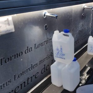 Puede incluir: Varios bidones de agua de pl&aacute;stico transparente con tapas azules est&aacute;n apilados cerca de una fuente. Los bidones tienen un asa y una etiqueta con la palabra "Lourdes" y una imagen religiosa. La fuente tiene una fachada de piedra gris oscura con texto grabado.