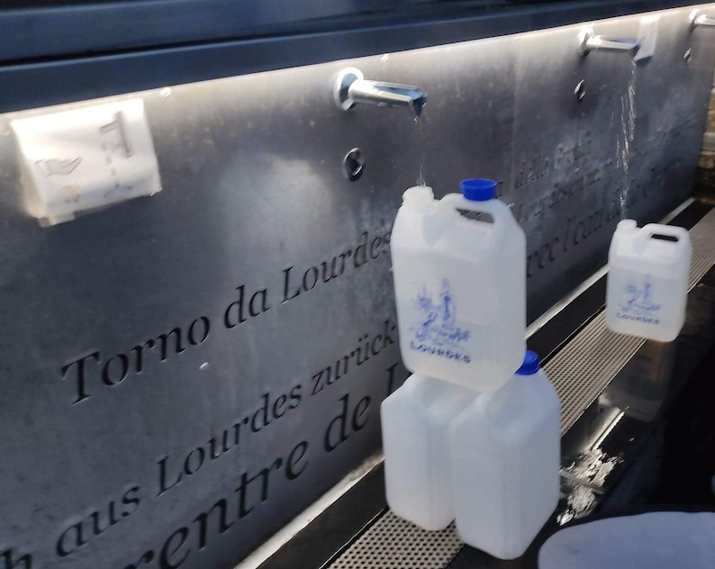 Puede incluir: Varios bidones de agua de pl&aacute;stico transparente con tapas azules est&aacute;n apilados cerca de una fuente. Los bidones tienen un asa y una etiqueta con la palabra "Lourdes" y una imagen religiosa. La fuente tiene una fachada de piedra gris oscura con texto grabado.