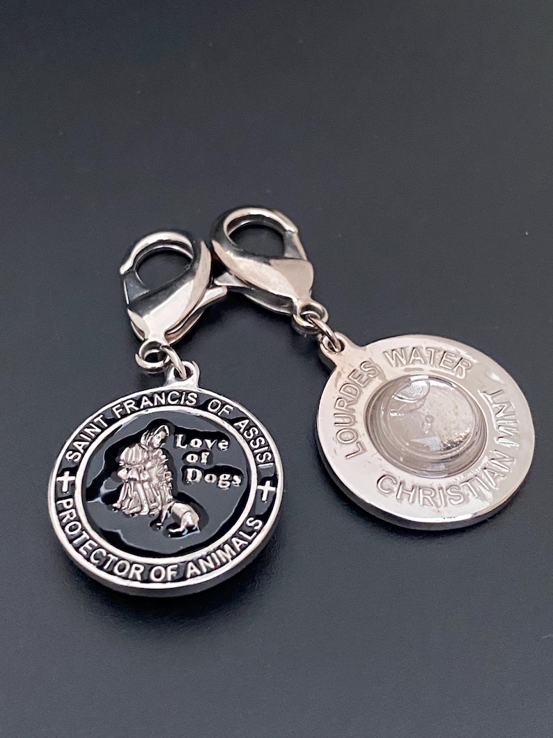 Puede incluir: Dos colgantes plateados con forma de perro. Un colgante presenta el texto "San Francisco de As&iacute;s, Protector de Animales, Amor de Perros". El otro colgante tiene el texto "Lourdes Water Christian Mint."